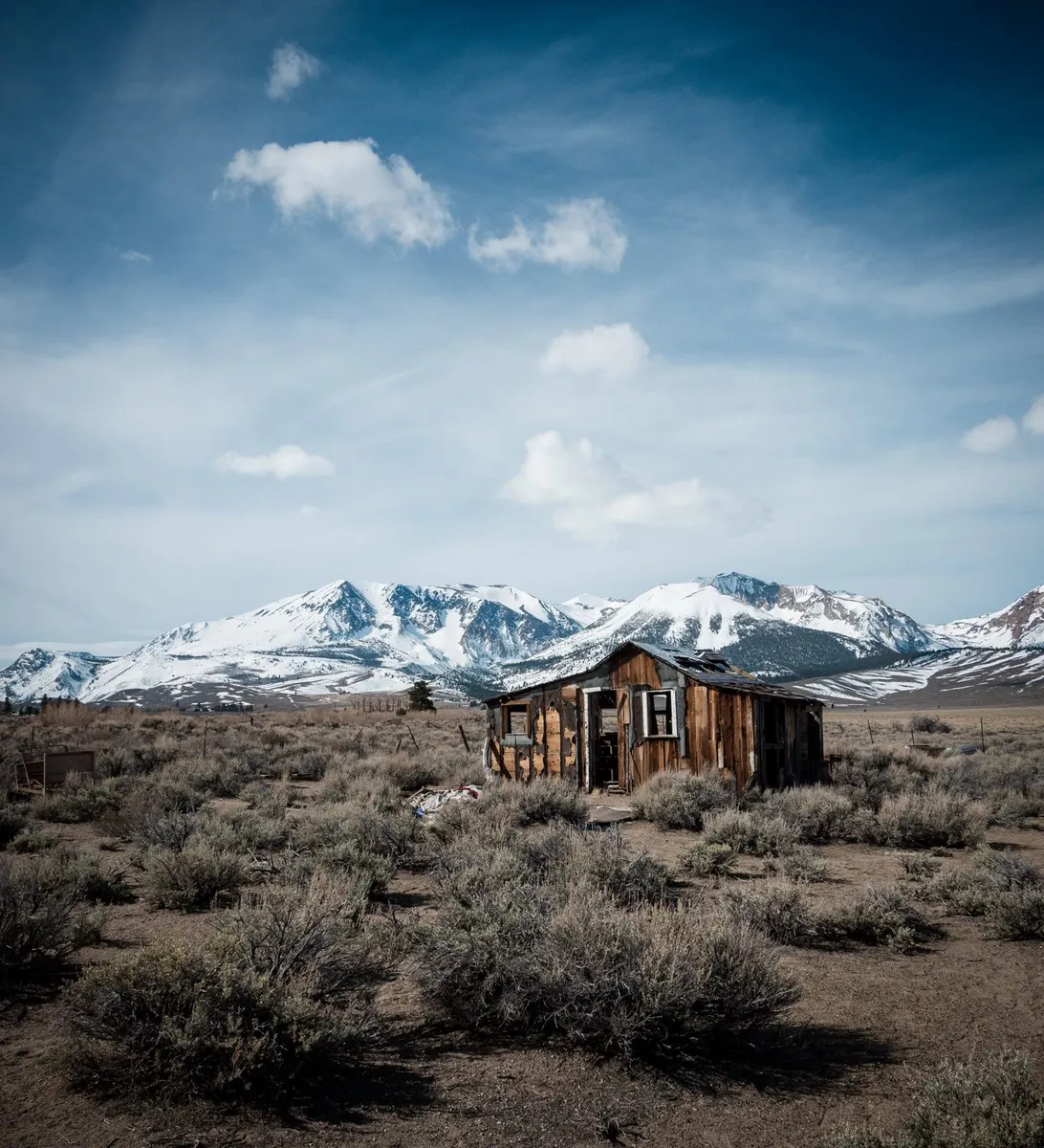 A weathered wooden cabin in sagebrush below snow-covered mountains and a blue sky with clouds