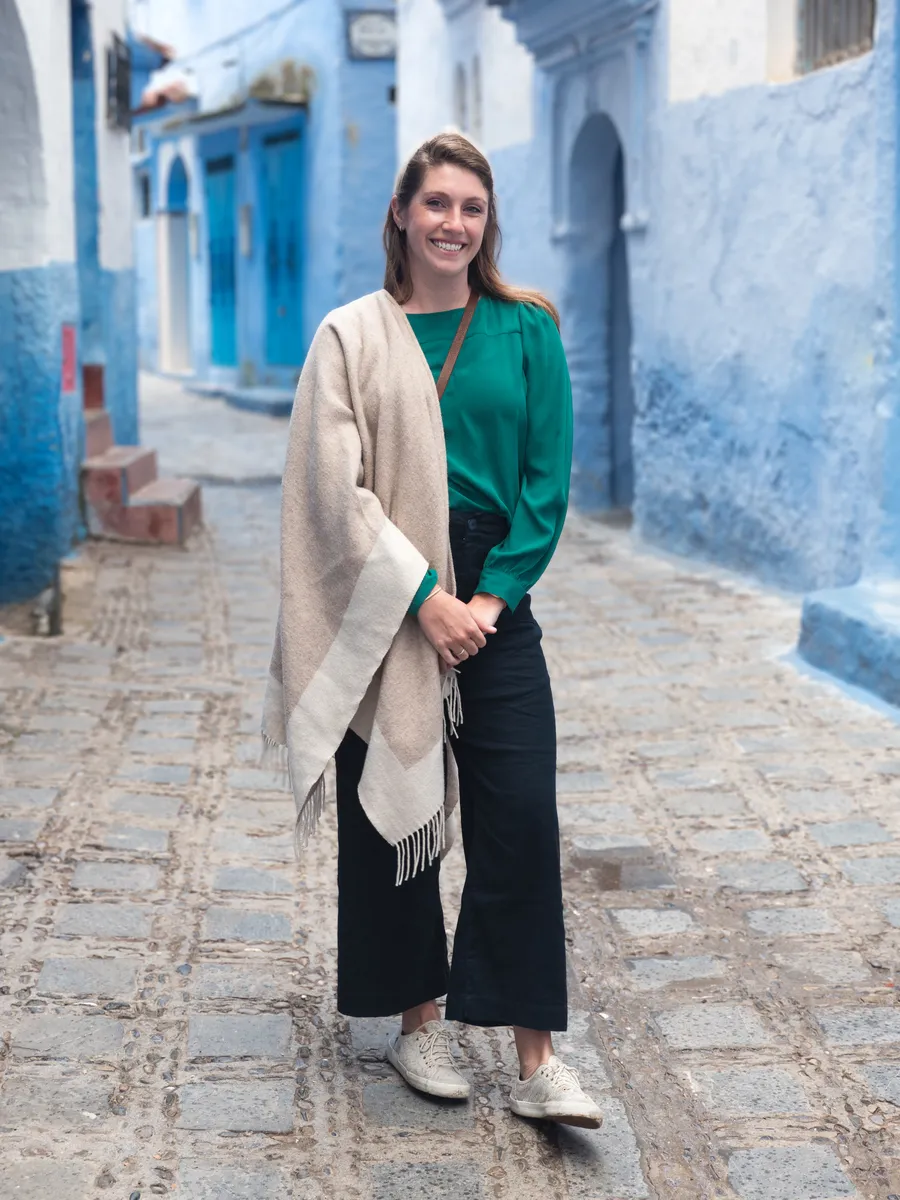 A smiling woman in a green top and cream shawl standing in a narrow blue-painted street