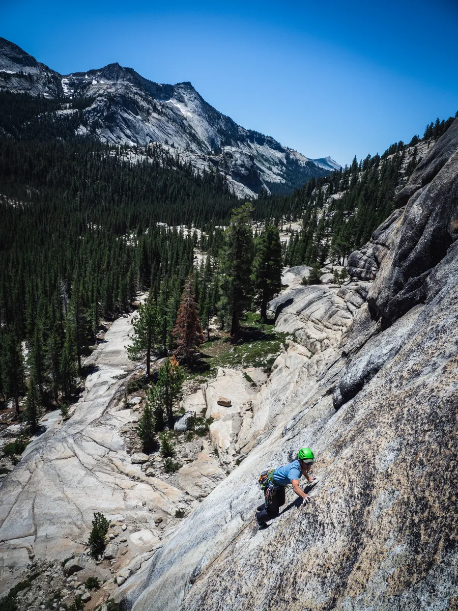 A climber in a green helmet on a granite slab above a pine-filled canyon