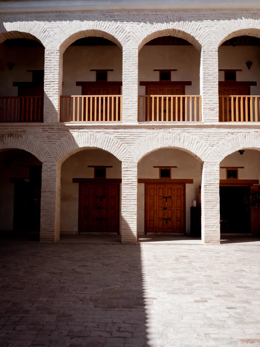 A two-story courtyard of brick arches and carved wooden doors, half in shade