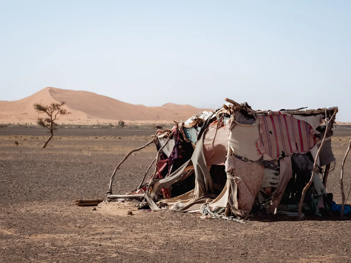 A patched tent of fabric and branches on a gravel plain, orange dunes on the horizon