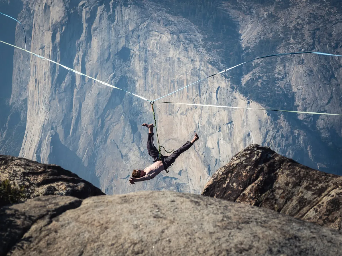 A highliner hanging from a line strung between rock outcrops above a huge granite wall