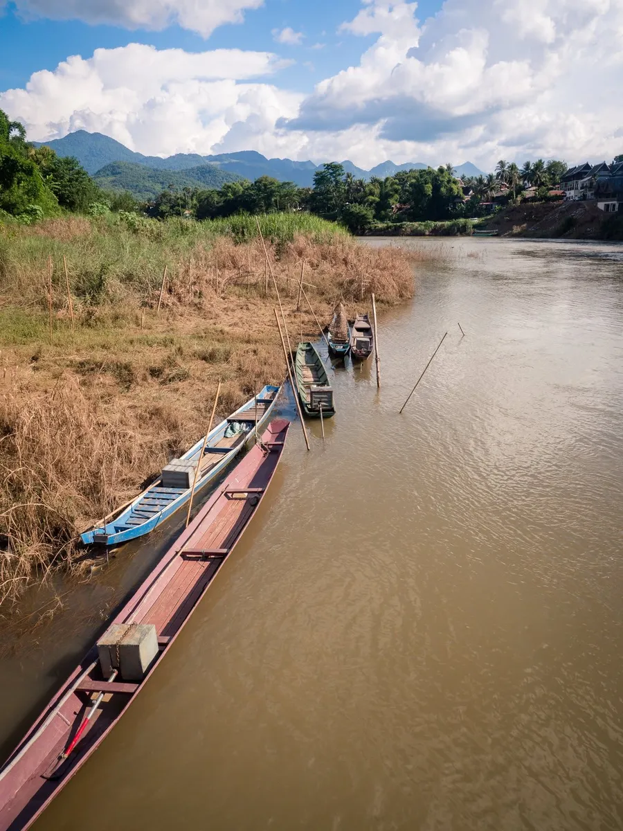 Long wooden boats moored along a muddy river bank below green hills