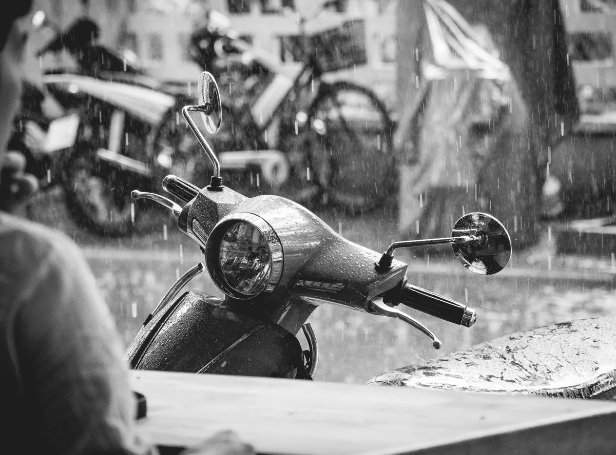 Black-and-white photo of a moped's headlight and mirrors in pouring rain
